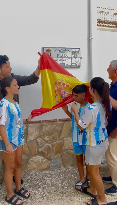 Jóvenes con camisetas blanquiazules sostienen una bandera de España ante el rótulo de la plaza Málaga C.F.