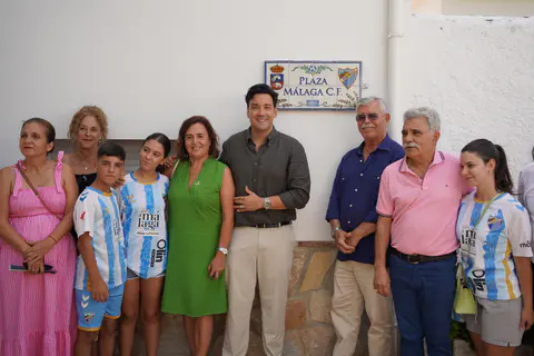 Grupo de asistentes posando bajo el rótulo cerámico de la plaza Málaga C.F. en Alfarnatejo.